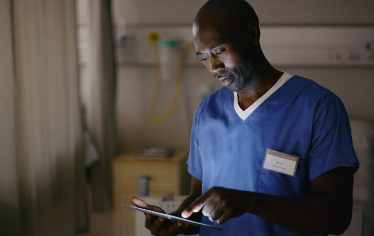 Male nurse in blue scrubs focused on a mobile device screen in a clinical setting.