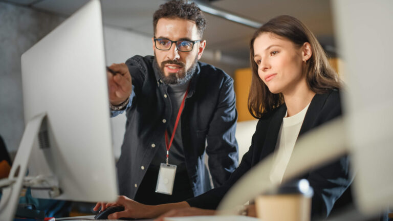 Two office colleagues, a man and a woman, discussing work while looking at a computer monitor.