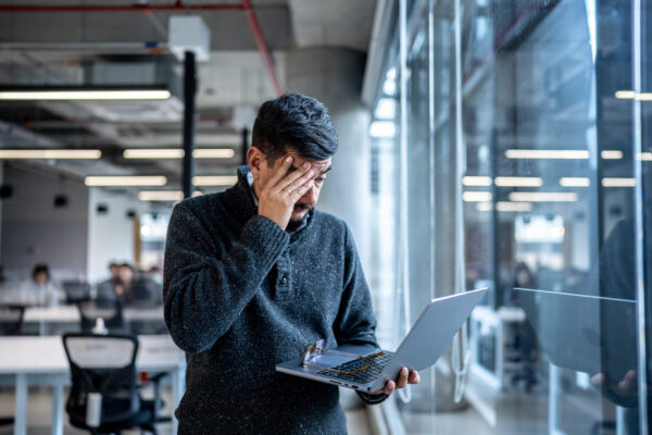 A man in an office looking overwhelmed while holding a laptop and touching his forehead.