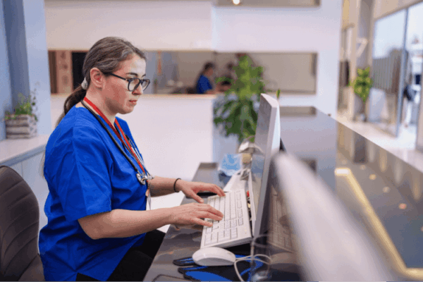 A nurse in blue scrubs working on a computer at a hospital reception desk.
