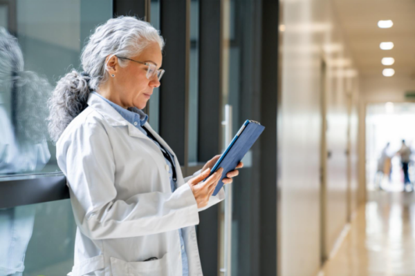 Female healthcare professional in a lab coat reviewing data on a digital tablet in a bright hallway.